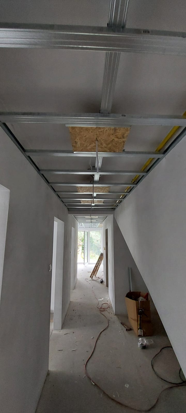 Korytarz w trakcie remontu: surowy tynk na ścianach, metalowa konstrukcja sufitu podwieszanego z widocznymi instalacjami. W tle drabina i narzędzia.