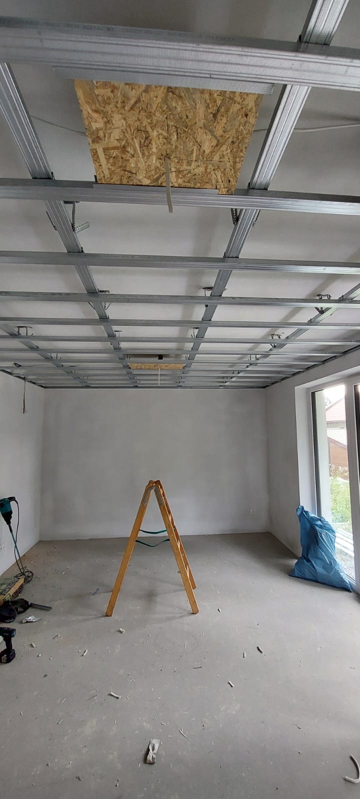 Montaż sufitu podwieszanego z metalową konstrukcją i płytą OSB w otworze. W tle ściany po gładzi, drabina i narzędzia. Etap prac wykończeniowych.