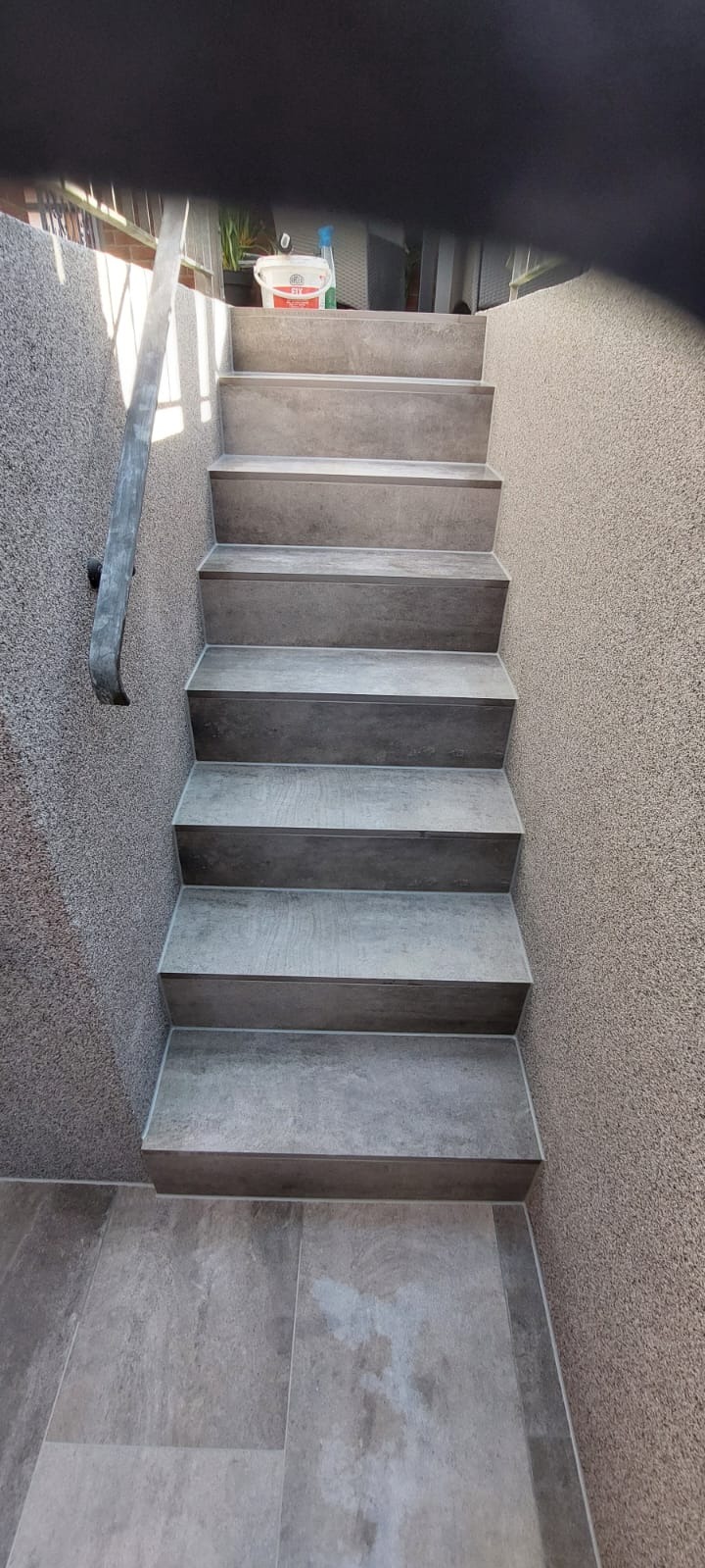 Wykończone schody z szarymi płytkami imitującymi drewno, prowadzące w dół. Ściany z jasnoszarego tynku strukturalnego. Widoczna poręcz i wiadro z farbą na górze schodów.