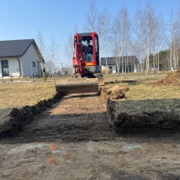 Z Gruntu Dobrze Szymon Szczerepa Roboty Ziemne - Czerwona mini koparka w trakcie wykopu pod fundamenty w Lubartowie. Widoczny operator i świeży wykop w ziemi. W tle dom i drzewa.