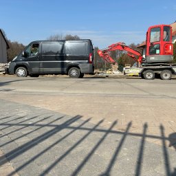 Z Gruntu Dobrze Szymon Szczerepa Roboty Ziemne - Czarny bus z przyczepą, na której stoi czerwona mini koparka Bobcat E26, zaparkowany na ulicy w słoneczny dzień. W tle domy i drzewa.