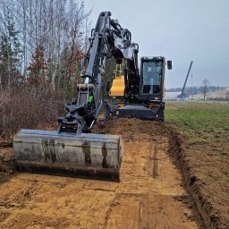 Z Gruntu Dobrze Szymon Szczerepa Roboty Ziemne - Żółto-czarna koparka kołowa Volvo z łyżką, wyrównująca teren na polu. W tle drzewa i dźwig. Świeżo wyrównana, wilgotna ziemia.
