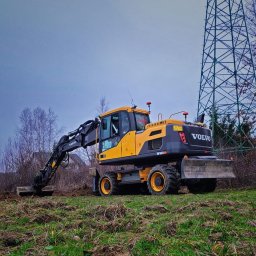 Z Gruntu Dobrze Szymon Szczerepa Roboty Ziemne - Żółta koparka kołowa Volvo EW160D w trakcie prac ziemnych na nierównym terenie, z widoczną linią wysokiego napięcia w tle.