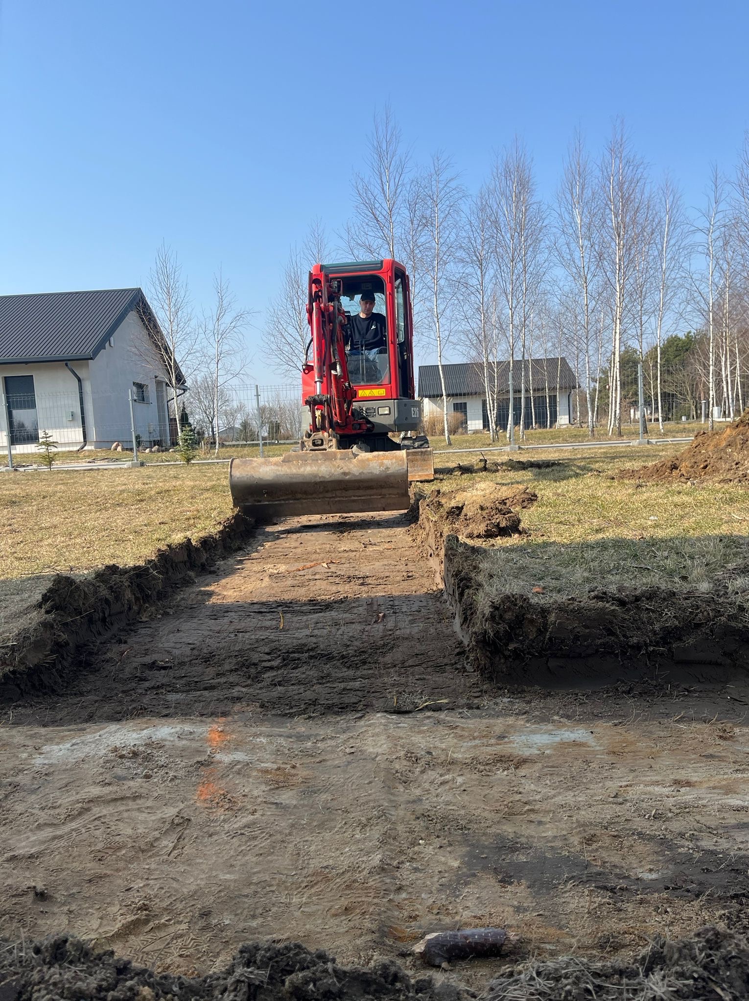 Czerwona mini koparka w trakcie wykopu pod fundamenty w Lubartowie. Widoczny operator i świeży wykop w ziemi. W tle dom i drzewa.
