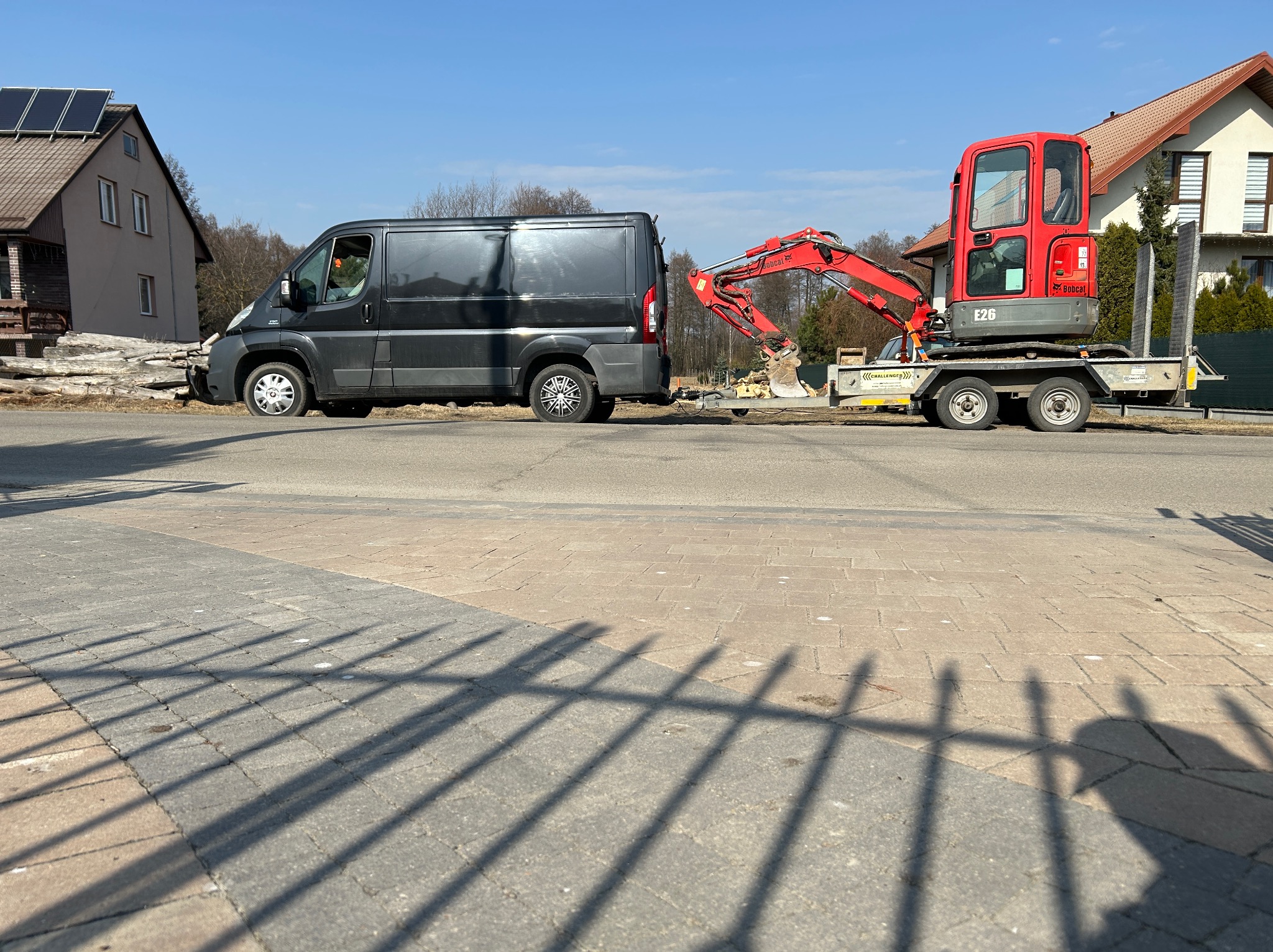 Czarny bus z przyczepą, na której stoi czerwona mini koparka Bobcat E26, zaparkowany na ulicy w słoneczny dzień. W tle domy i drzewa.