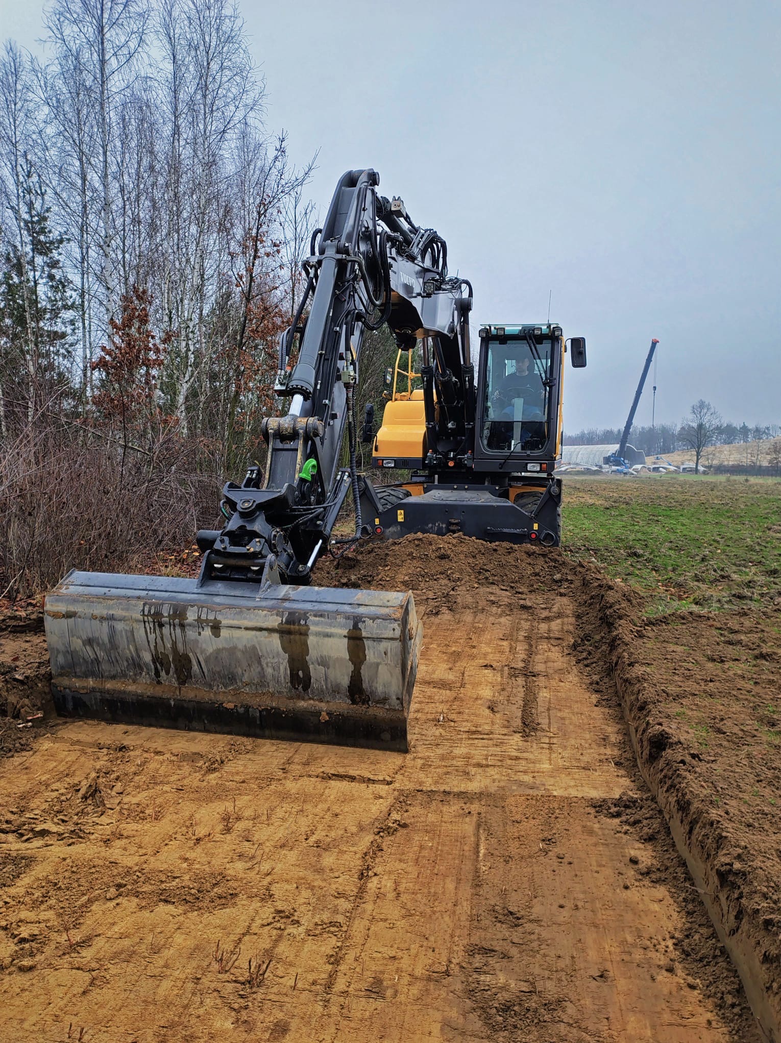 Żółto-czarna koparka kołowa Volvo z łyżką, wyrównująca teren na polu. W tle drzewa i dźwig. Świeżo wyrównana, wilgotna ziemia.