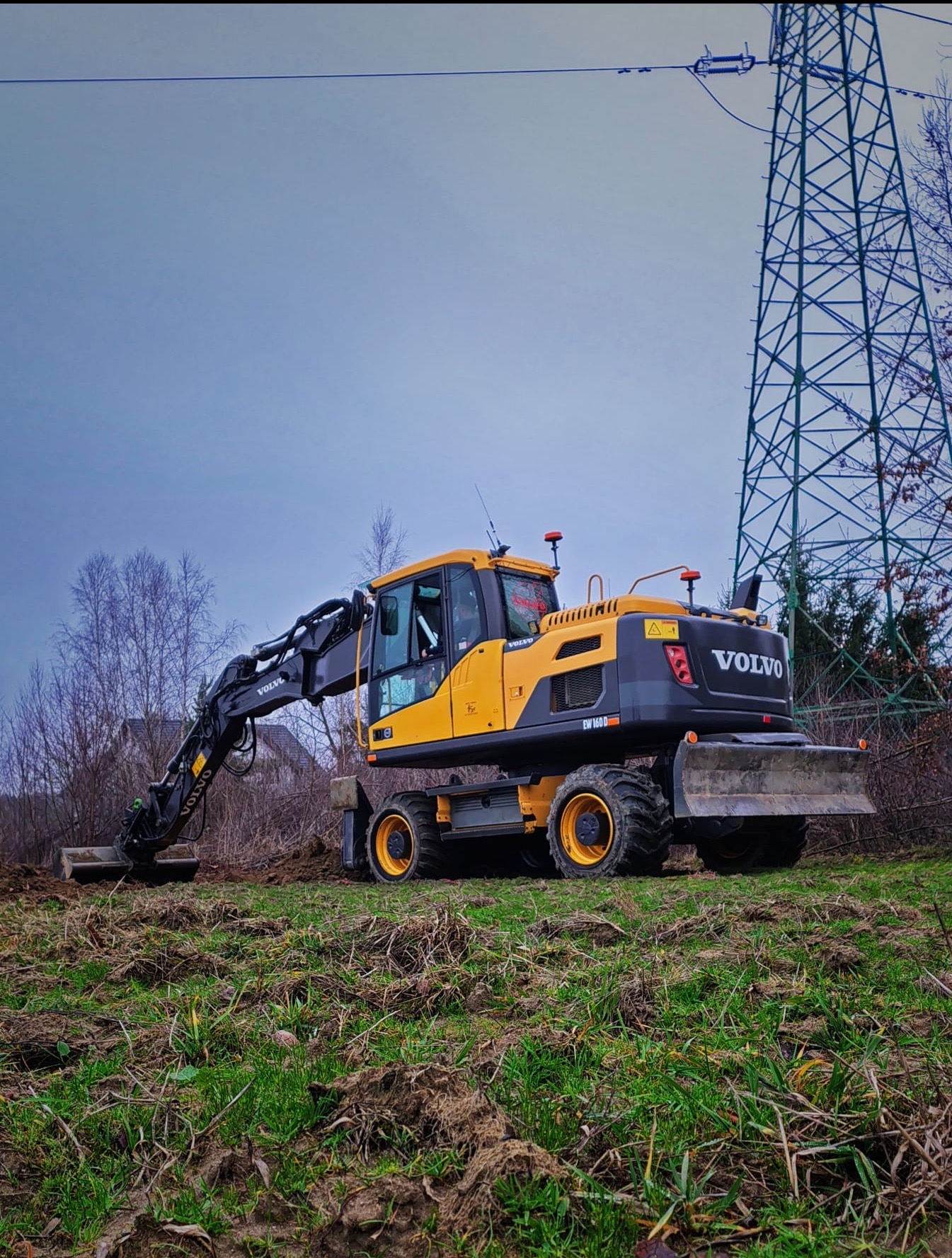 Żółta koparka kołowa Volvo EW160D w trakcie prac ziemnych na nierównym terenie, z widoczną linią wysokiego napięcia w tle.