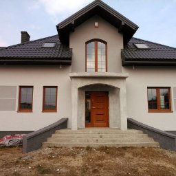 DB-est - Elewacja domu jednorodzinnego w odcieniach szarości z brązowymi oknami i drzwiami wejściowymi. Widoczne schody i fragment ogrodu. Dom z poddaszem użytkowym i ciemną dachówką.