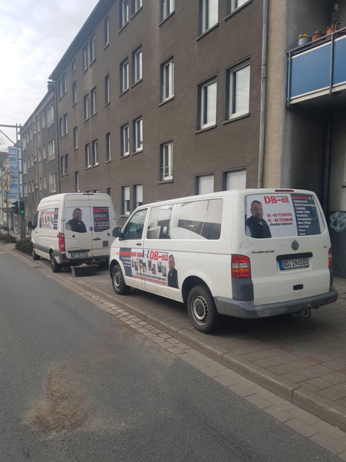 Dwa białe busy firmy remontowej zaparkowane przy budynku mieszkalnym. Na busach reklama z ofertą usług i zdjęciem pracownika. Tablice rejestracyjne niemieckie.