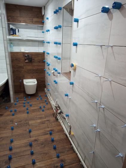 Układanie płytek w łazience: brązowe płytki imitujące drewno na podłodze, jasne płytki na ścianach, system poziomowania płytek z niebieskimi elementami.