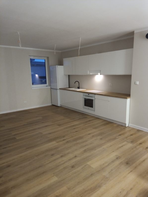 Jasne panele podłogowe w nowym mieszkaniu z białą zabudową kuchenną i sprzętem AGD. Widoczne niedokończone podłączenia elektryczne.