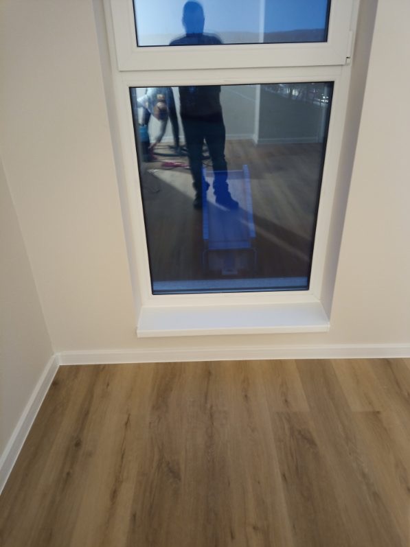 Jasne panele podłogowe w pokoju po remoncie, widoczne białe listwy przypodłogowe i okno z odbiciem osoby. Minimalistyczne wykończenie wnętrza.