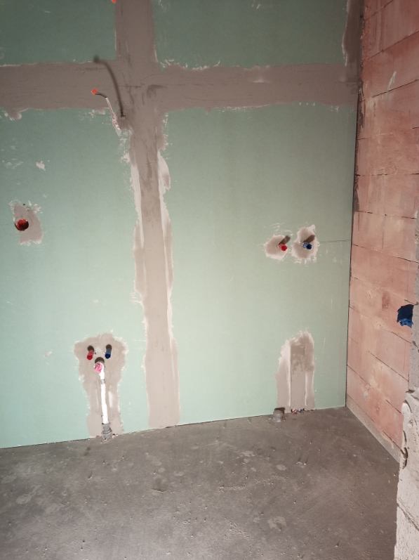 Ściana z płyt gipsowo-kartonowych po szpachlowaniu, widoczne instalacje wodne i elektryczne. Surowy stan wykończenia wnętrza, przygotowanie do dalszych prac.