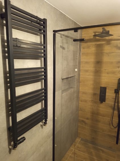 Nowoczesna łazienka z kabiną prysznicową typu walk-in, drewnopodobnymi płytkami i czarnym grzejnikiem drabinkowym na szarej ścianie.