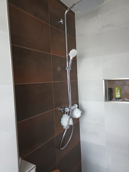 Nowoczesna kabina prysznicowa z brązowymi i białymi płytkami. Bateria natryskowa z deszczownicą i słuchawką. Wnęka w ścianie z kosmetykami. Minimalistyczny design.