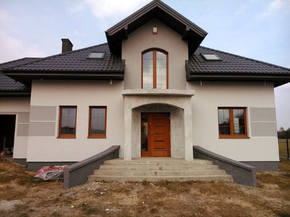 Elewacja domu jednorodzinnego w odcieniach szarości z brązowymi oknami i drzwiami wejściowymi. Widoczne schody i fragment ogrodu. Dom z poddaszem użytkowym i ciemną dachówką.