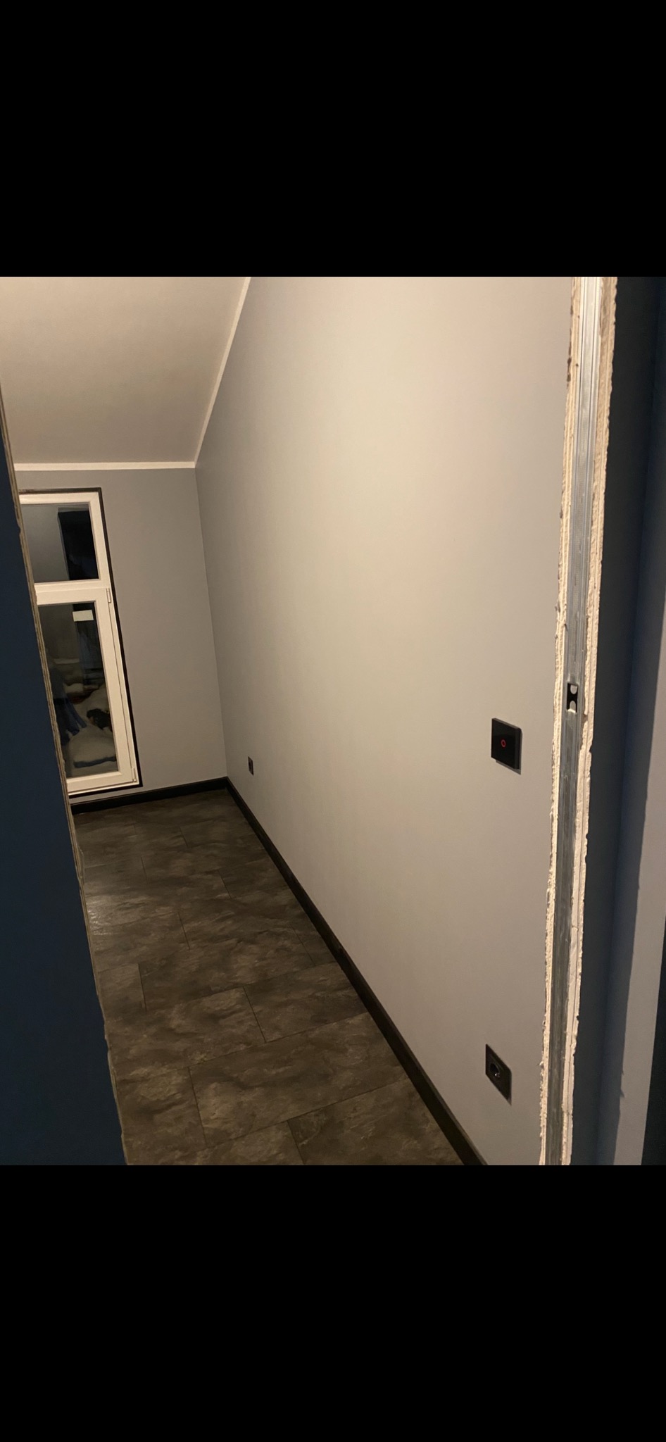 Korytarz po remoncie: szare ściany, ciemna podłoga z paneli, białe okno. Widoczne gniazdka i włączniki. Ościeżnica drzwiowa w trakcie montażu.