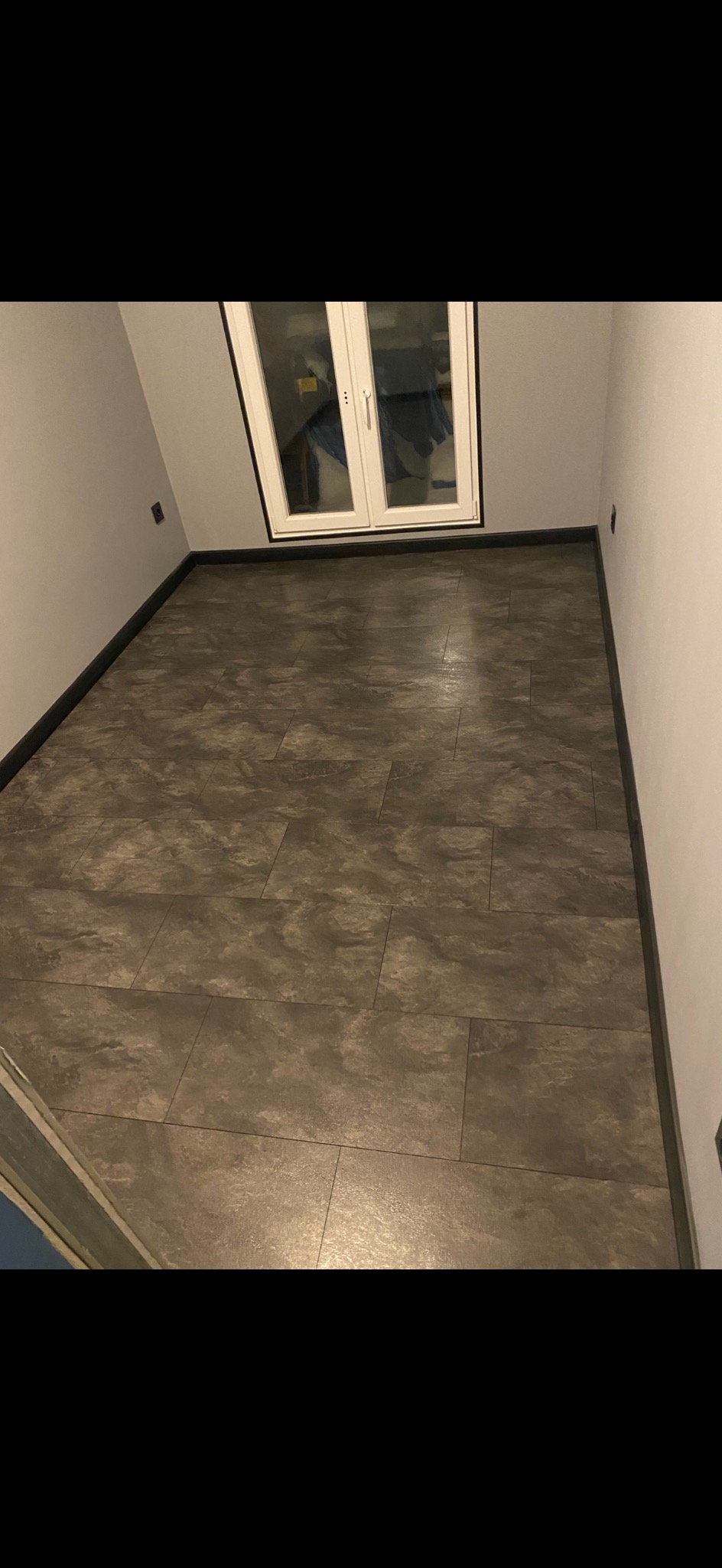 Ułożone szare panele podłogowe w wąskim pomieszczeniu z białymi drzwiami balkonowymi i czarnymi listwami przypodłogowymi. Widok z góry, lekki kąt.