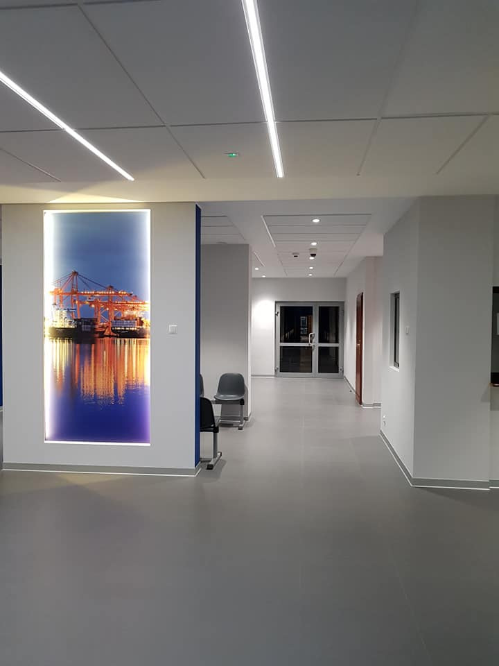 Nowoczesne oświetlenie LED w korytarzu biurowym w Gdyni. Podwieszany sufit z liniowymi lampami, na ścianie podświetlona grafika z motywem portowym.