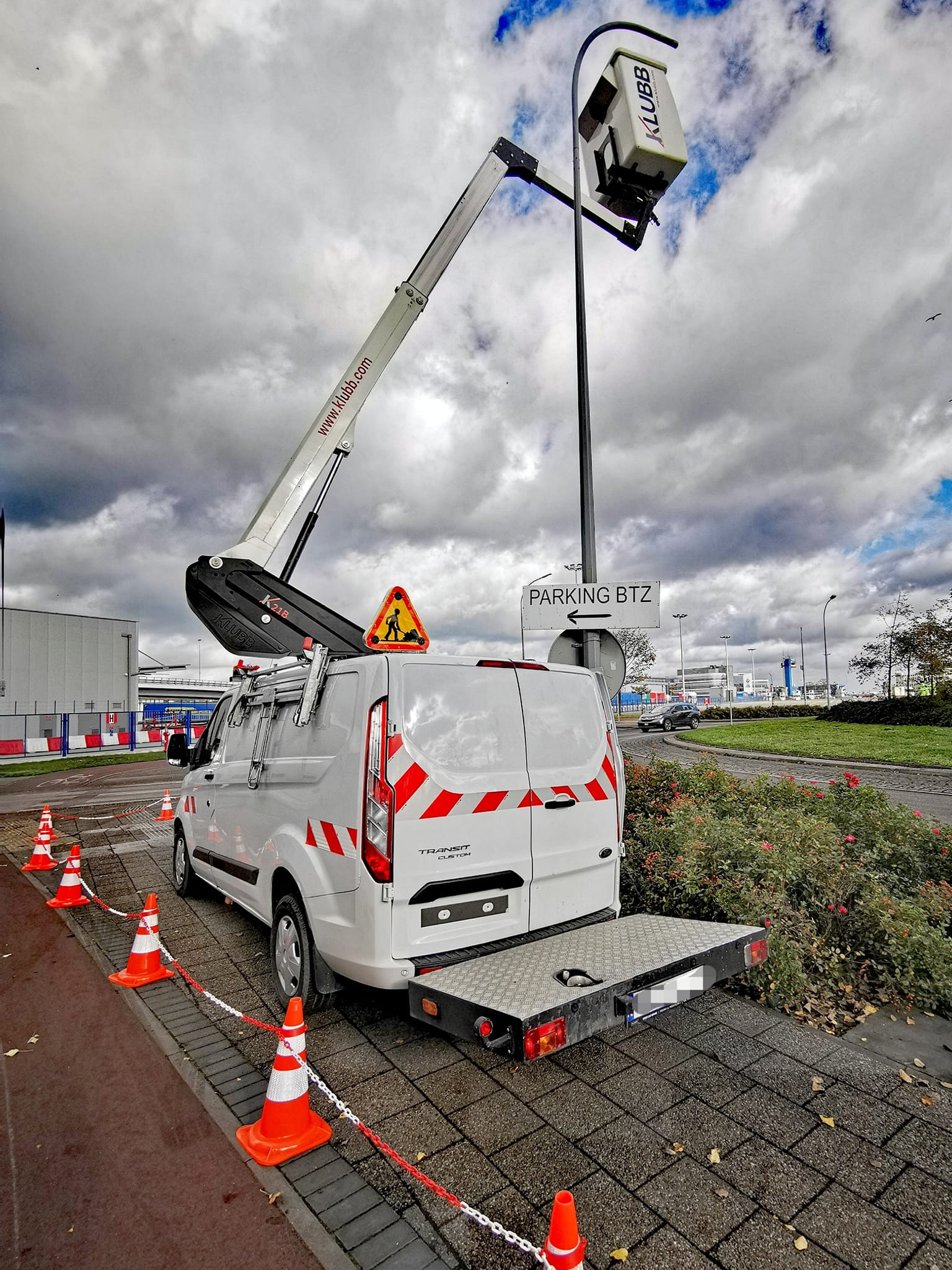 Biały furgon z podnośnikiem koszowym Klubb na tle pochmurnego nieba, otoczony pomarańczowymi pachołkami. Tablica 'Parking BTZ' w tle.