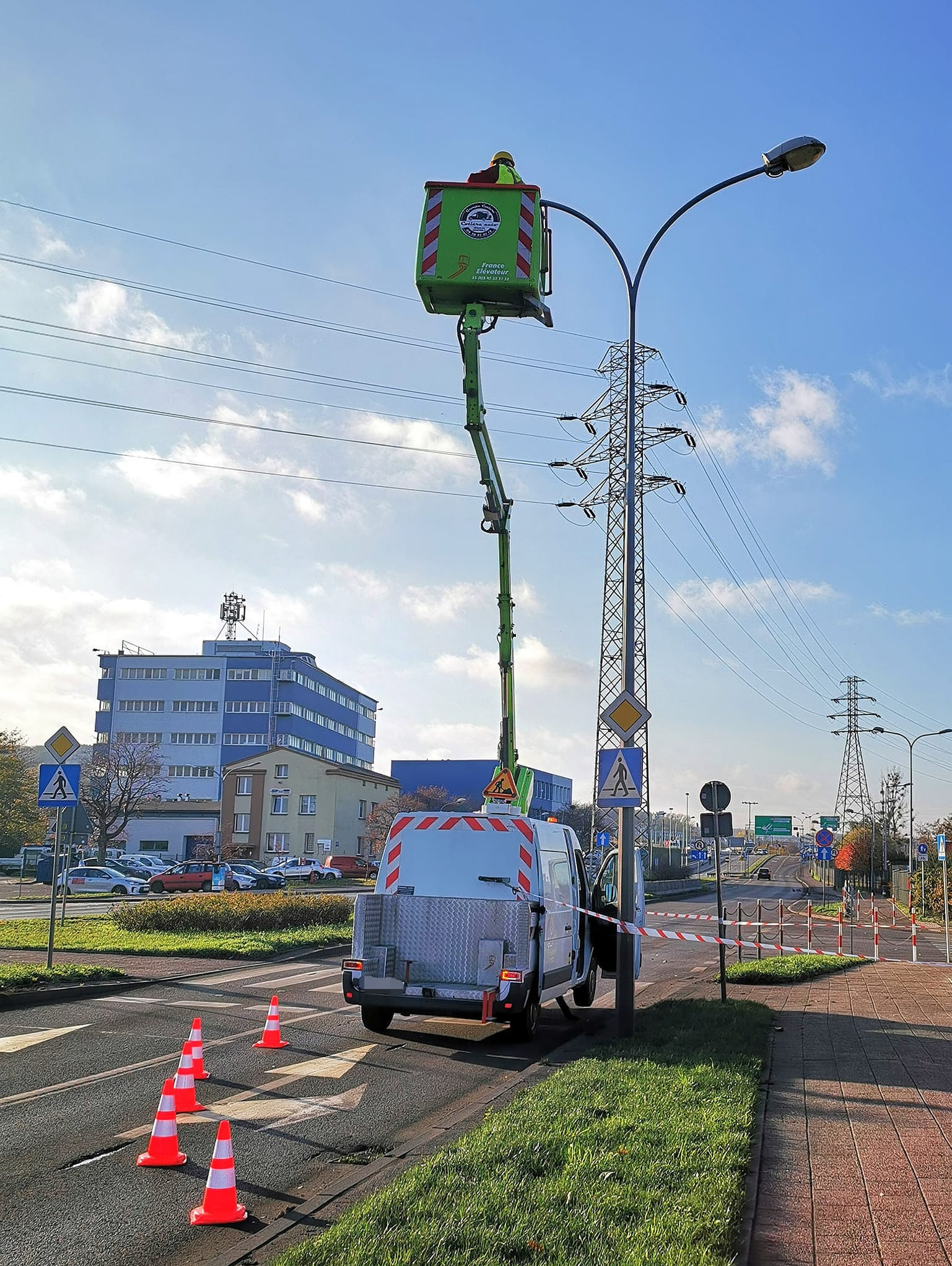 Pracownik w podnośniku koszowym naprawia oświetlenie uliczne, zabezpieczony obszar stożkami i taśmą ostrzegawczą. W tle budynek i słupy wysokiego napięcia.
