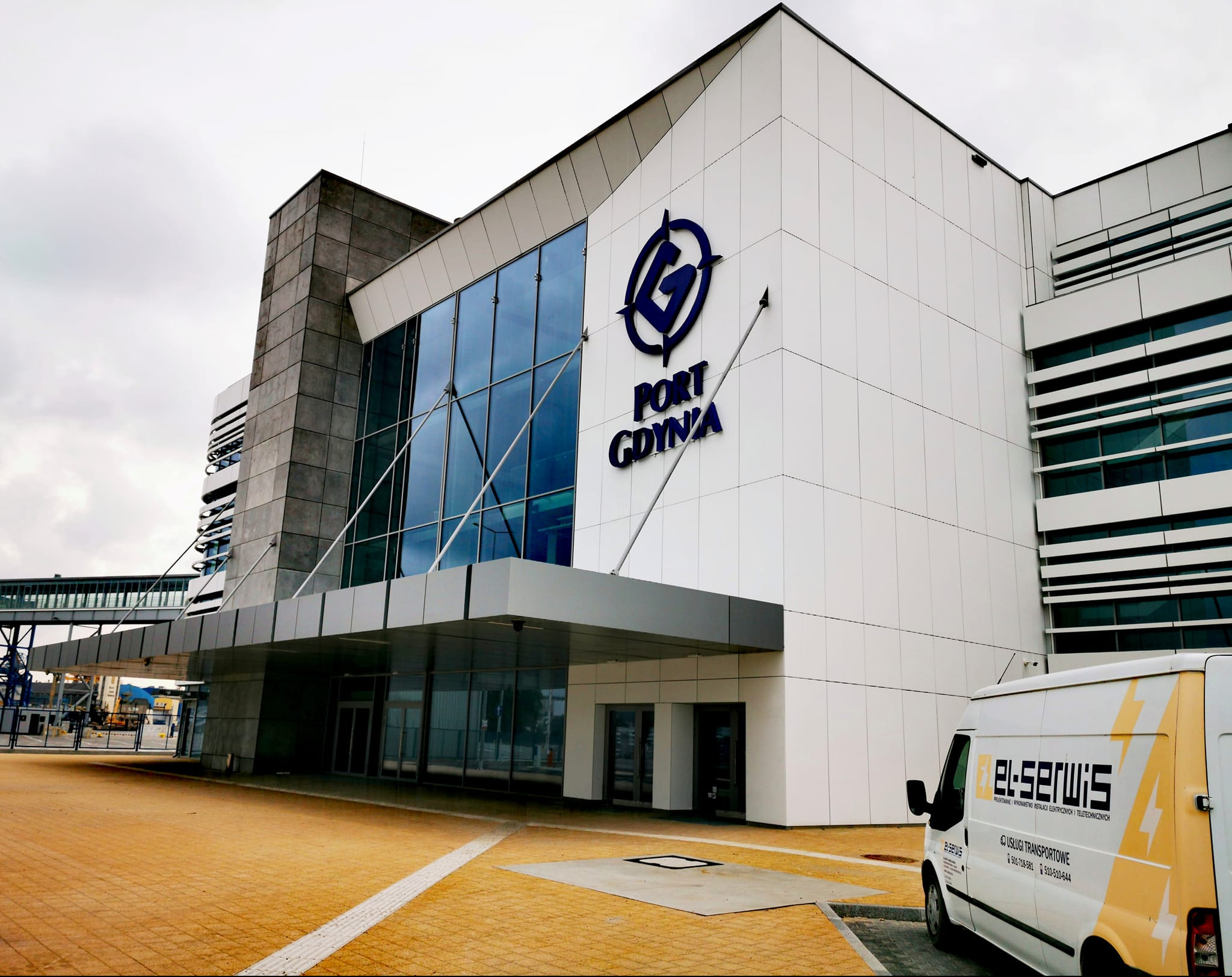 Budynek Portu Gdynia z logo i zaparkowanym vanem firmy elektrycznej z żółtymi akcentami i napisem 'el-serwis' na tle kostki brukowej.