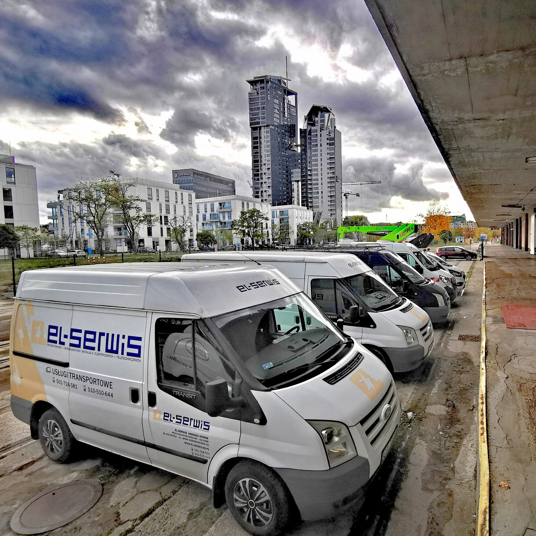 Flota białych samochodów dostawczych 'El-Serwis' na tle nowoczesnej architektury miejskiej i pochmurnego nieba. Widok z boku, perspektywa z dołu.