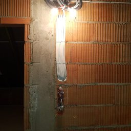 MARCIN WALAS - Instalacja elektryczna w trakcie montażu na ceglanej ścianie. Widoczne kable w peszlach, puszka elektryczna i wiązka przewodów. Oświetlenie budowlane.
