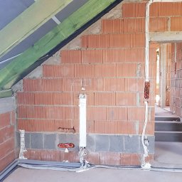 MARCIN WALAS - Wnętrze w trakcie prac instalacyjnych: widoczne przewody elektryczne prowadzone po ceglanej ścianie, szare rury wzdłuż podłogi i drewniana konstrukcja dachu.