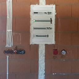 MARCIN WALAS - Rozdzielnica elektryczna z przewodami w ścianie z czerwonej cegły. Instalacja zabezpieczona pianką montażową. Widoczne kable i puszki instalacyjne.