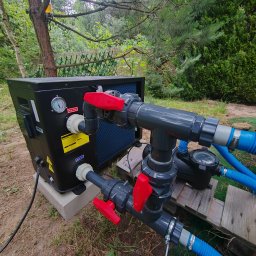 MARCIN WALAS - Czarna pompa ciepła z manometrem, podłączona do instalacji z rur PCV i zaworami z czerwonymi uchwytami, na zewnątrz, w otoczeniu zieleni.