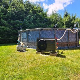 MARCIN WALAS - Zewnętrzna pompa ciepła do basenu ogrodowego, z podłączeniem do systemu filtracji. Widoczne węże, trawnik i drzewa iglaste w tle. Słoneczny dzień.