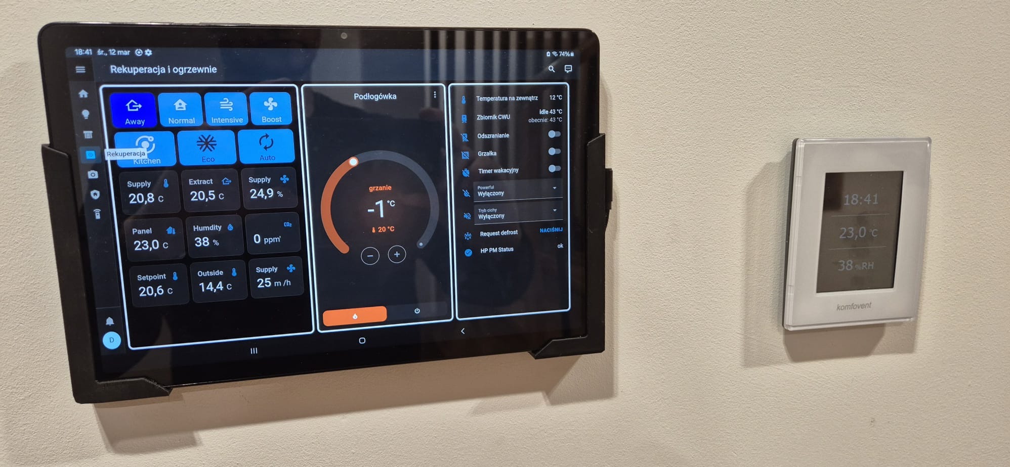 Tablet z aplikacją do sterowania rekuperacją i ogrzewaniem podłogowym, obok panel sterowania Komfovent. Widoczne parametry temperatury i wilgotności.