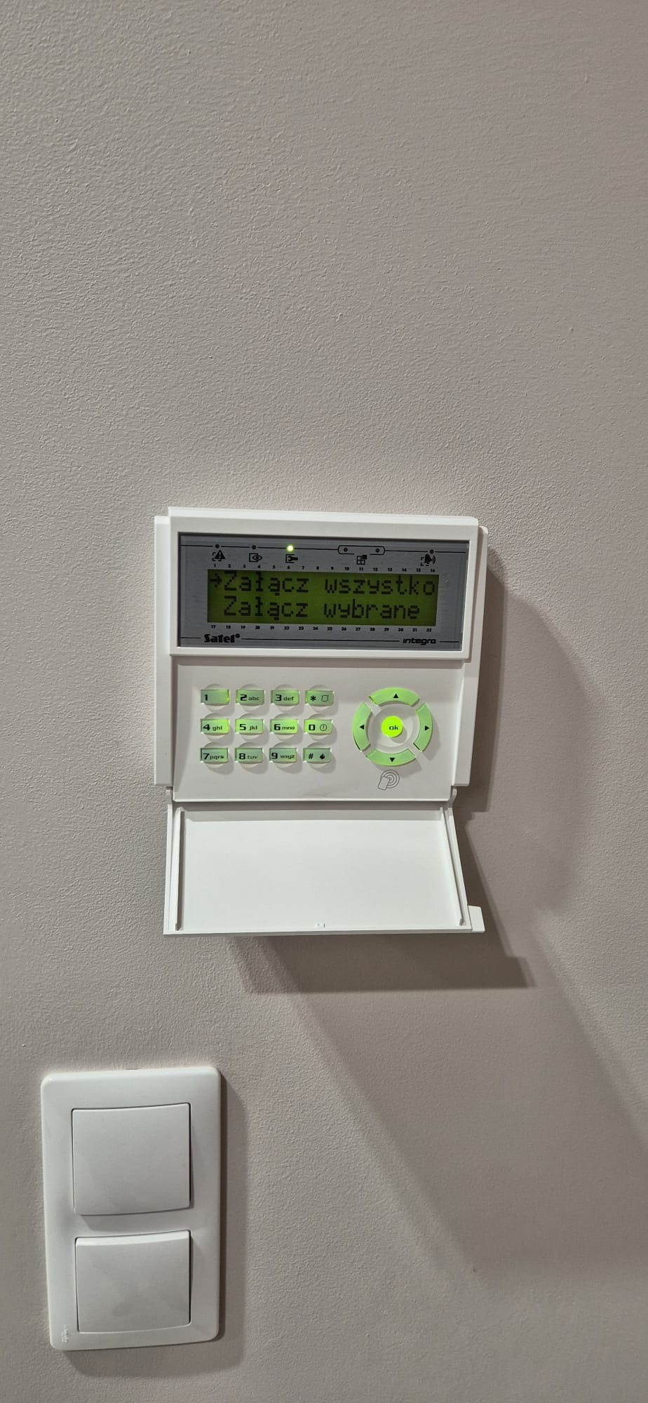 Panel sterowania alarmem Satel Integra na ścianie, z podświetlonym ekranem i klawiaturą. Obok włącznik światła. Widoczne cienie.