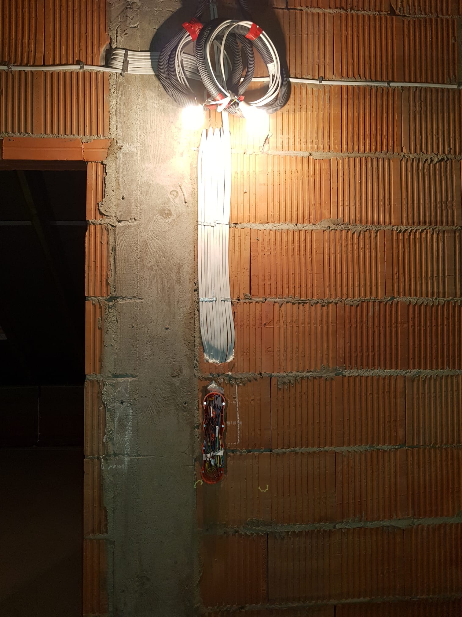 Instalacja elektryczna w trakcie montażu na ceglanej ścianie. Widoczne kable w peszlach, puszka elektryczna i wiązka przewodów. Oświetlenie budowlane.