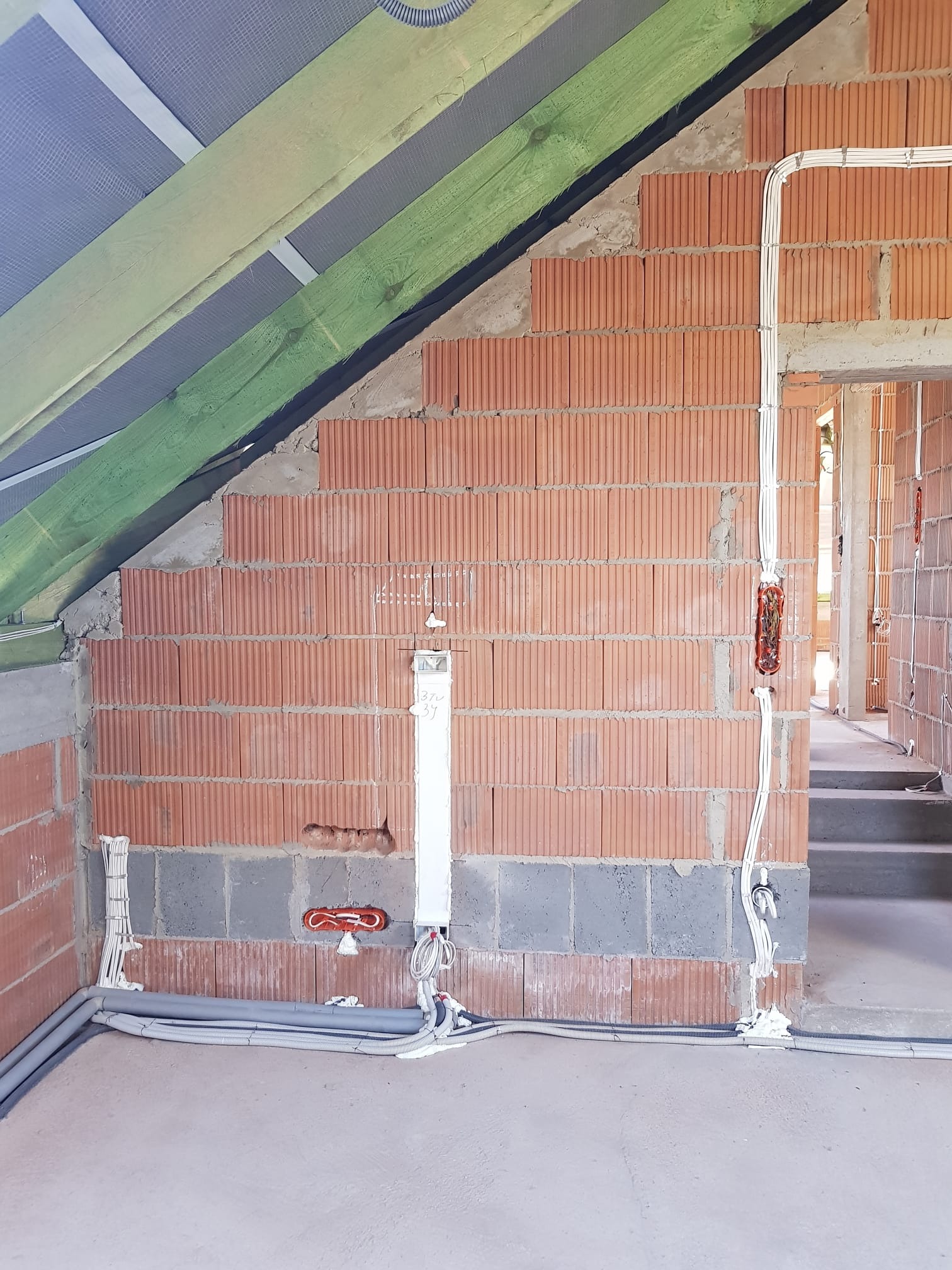 Wnętrze w trakcie prac instalacyjnych: widoczne przewody elektryczne prowadzone po ceglanej ścianie, szare rury wzdłuż podłogi i drewniana konstrukcja dachu.