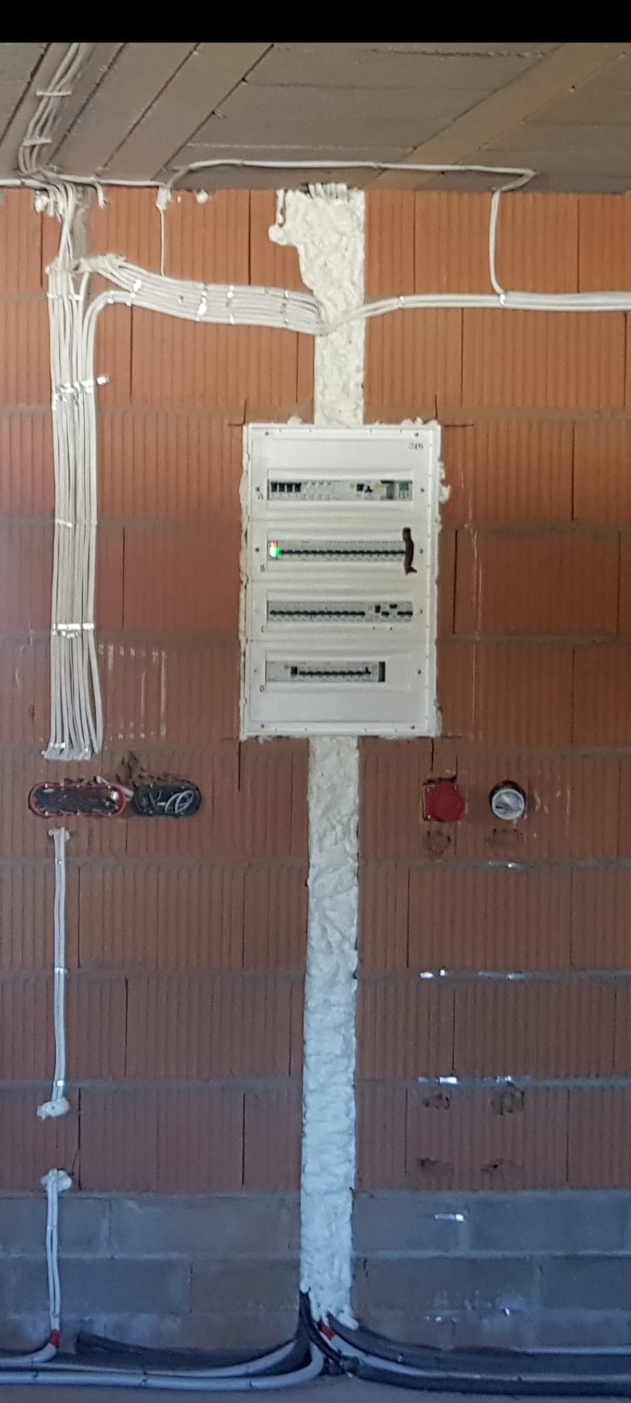 Rozdzielnica elektryczna z przewodami w ścianie z czerwonej cegły. Instalacja zabezpieczona pianką montażową. Widoczne kable i puszki instalacyjne.
