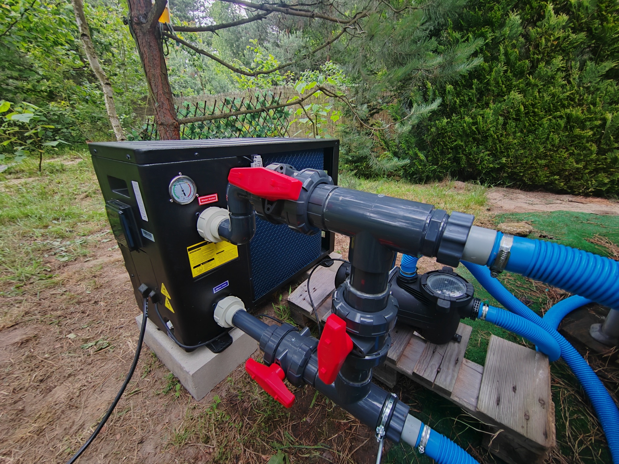 Czarna pompa ciepła z manometrem, podłączona do instalacji z rur PCV i zaworami z czerwonymi uchwytami, na zewnątrz, w otoczeniu zieleni.