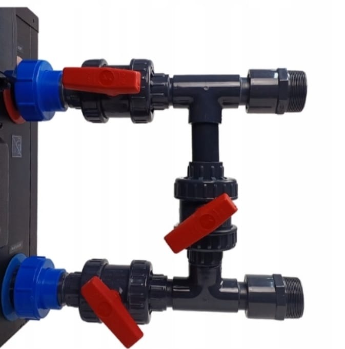 Szare rury z czerwonymi zaworami i niebieskimi złączkami, tworzące symetryczny układ na białym tle, przymocowane do ciemnej skrzynki. Widoczne gwintowane końcówki.