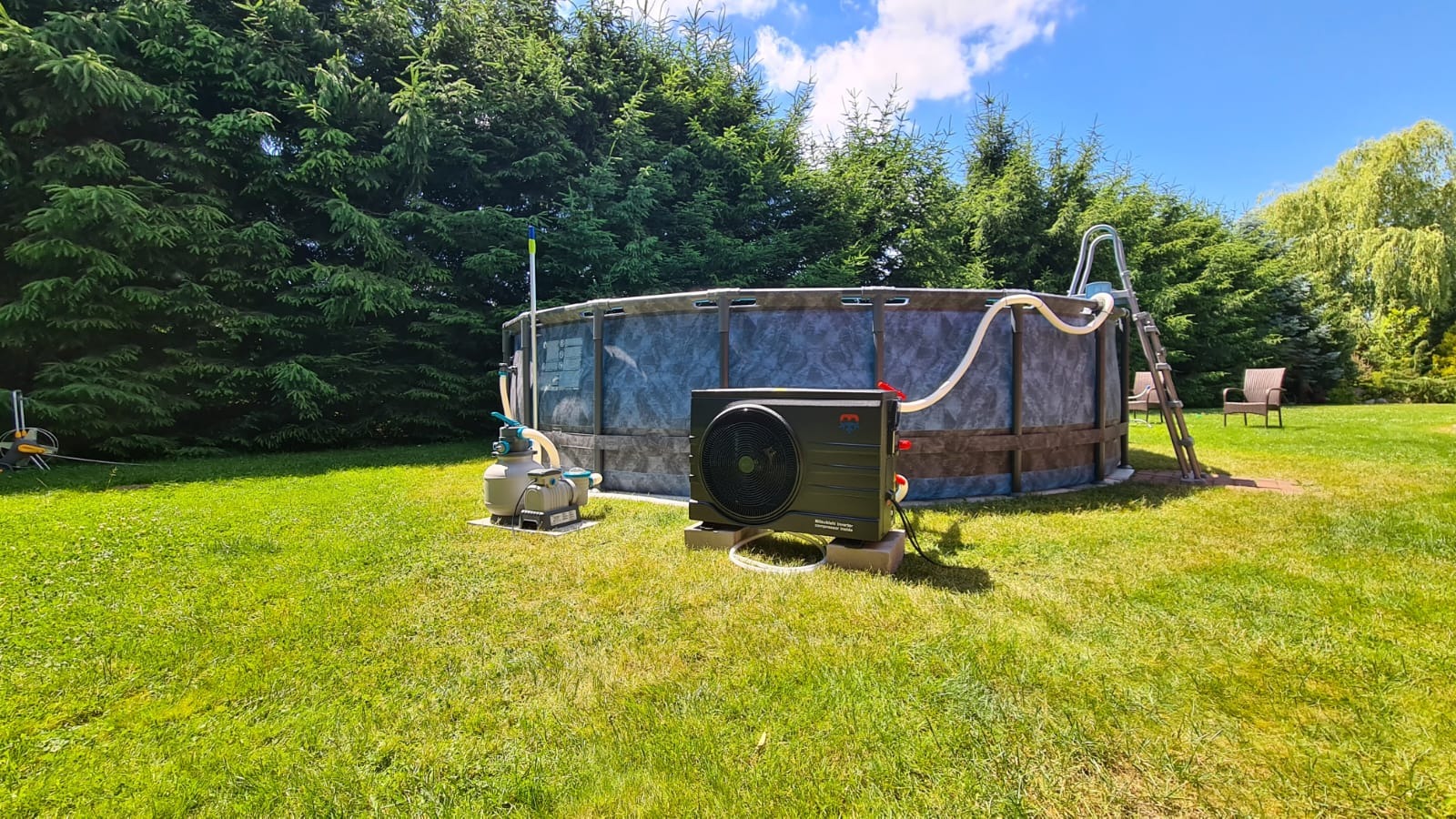Zewnętrzna pompa ciepła do basenu ogrodowego, z podłączeniem do systemu filtracji. Widoczne węże, trawnik i drzewa iglaste w tle. Słoneczny dzień.