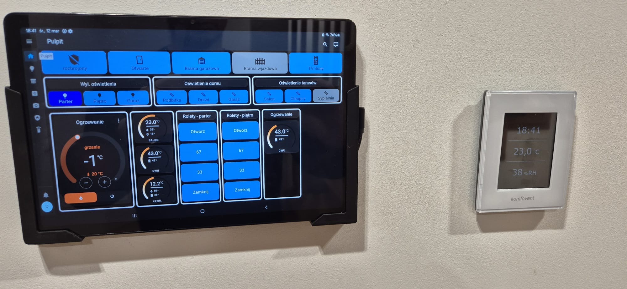 Tablet z panelem sterowania inteligentnym domem, obok panel ścienny Komfovent. Widoczne ustawienia ogrzewania, rolet, oświetlenia. Nowoczesne rozwiązania dla domu.