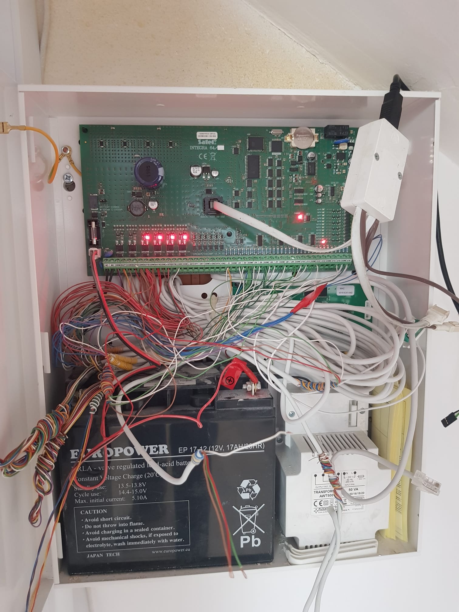 Otwarta skrzynka z elektroniką alarmową: płyta główna, akumulator Europower, transformator Pulsar, liczne przewody. Widoczne diody LED i złącza. Instalacja w białej obudowie.
