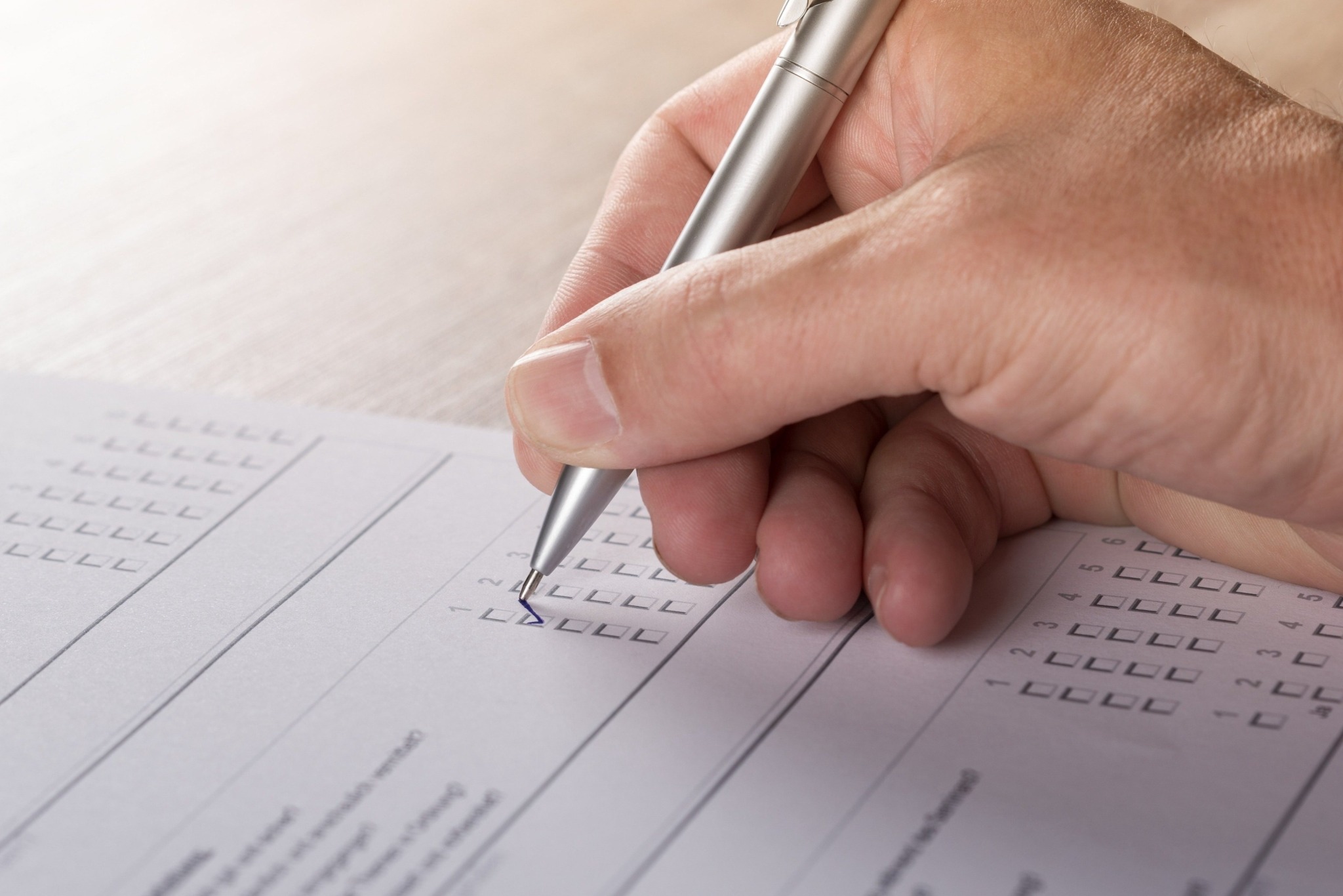 Ręka wypełnia ankietę srebrnym długopisem, zaznaczając kwadratowe pola wyboru. Jasne tło, delikatne światło podkreśla detale formularza.
