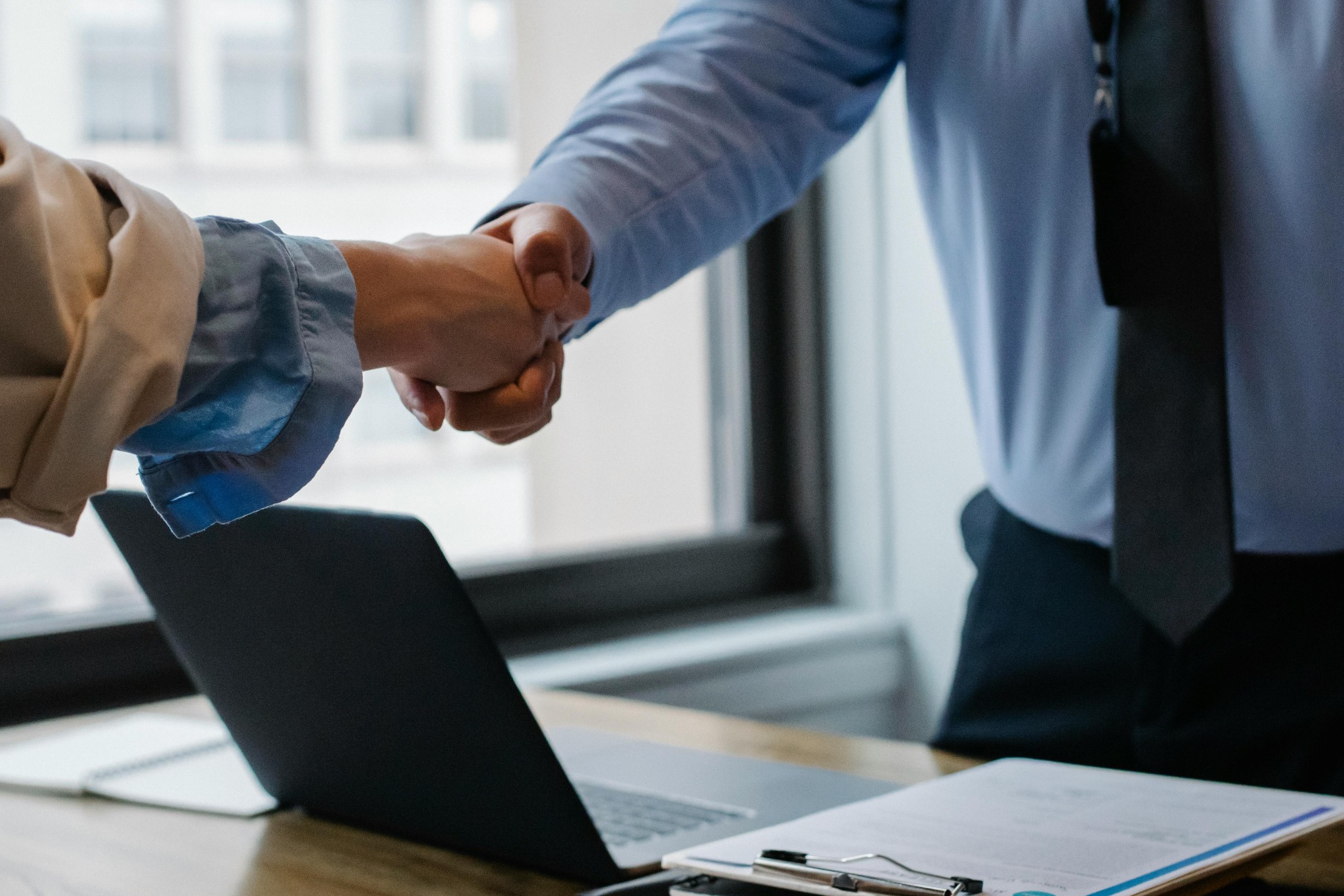 Uścisk dłoni nad laptopem i dokumentami. Symbol zawarcia umowy lub partnerstwa biznesowego. Widoczne okno w tle. Ujęcie z bliska, skupione na dłoniach.