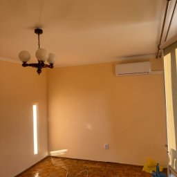 Rafal Kubiak - Pomarańczowy pokój po malowaniu z parkietem, lampą i klimatyzacją. Widoczny kabel i narzędzia, sugerujące prace wykończeniowe.