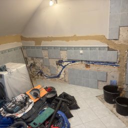 Fragment łazienki w trakcie remontu: częściowo położone szare płytki, widoczne instalacje, narzędzia i pralka zabezpieczona folią. Ujęcie z bliska, wnętrze pod skosem.