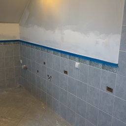 Łazienka w trakcie prac: szare płytki na ścianach do połowy wysokości, zabezpieczona folią podłoga, ściany przygotowane do malowania, widoczne punkty instalacji.