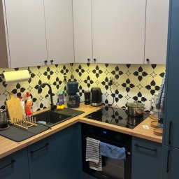 Nowoczesna kuchnia z białymi szafkami górnymi i niebieskimi dolnymi, drewnianym blatem i czarną armaturą. Na ścianie dekoracyjne płytki w kwiatowy wzór.