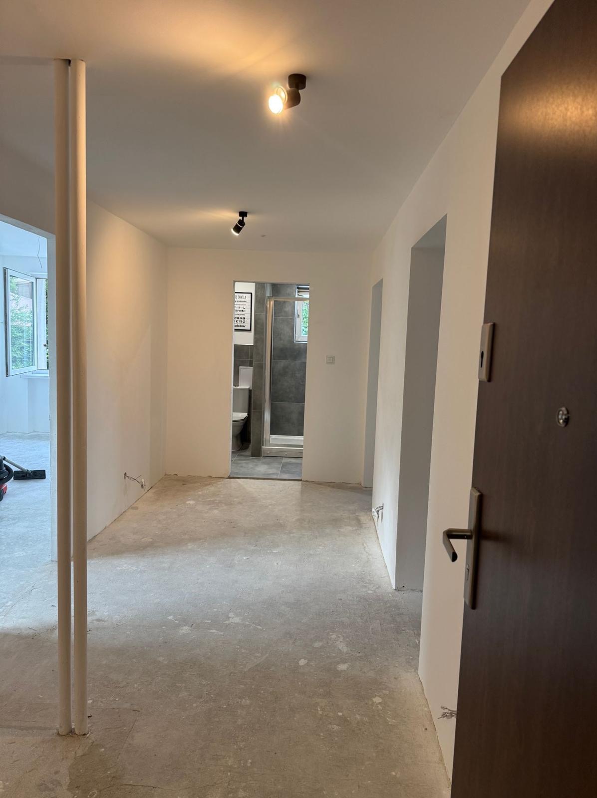 Wnętrze mieszkania w Krakowie po malowaniu ścian na biało. Widoczny surowy beton na podłodze, drzwi wejściowe i wejście do łazienki z kabiną prysznicową.