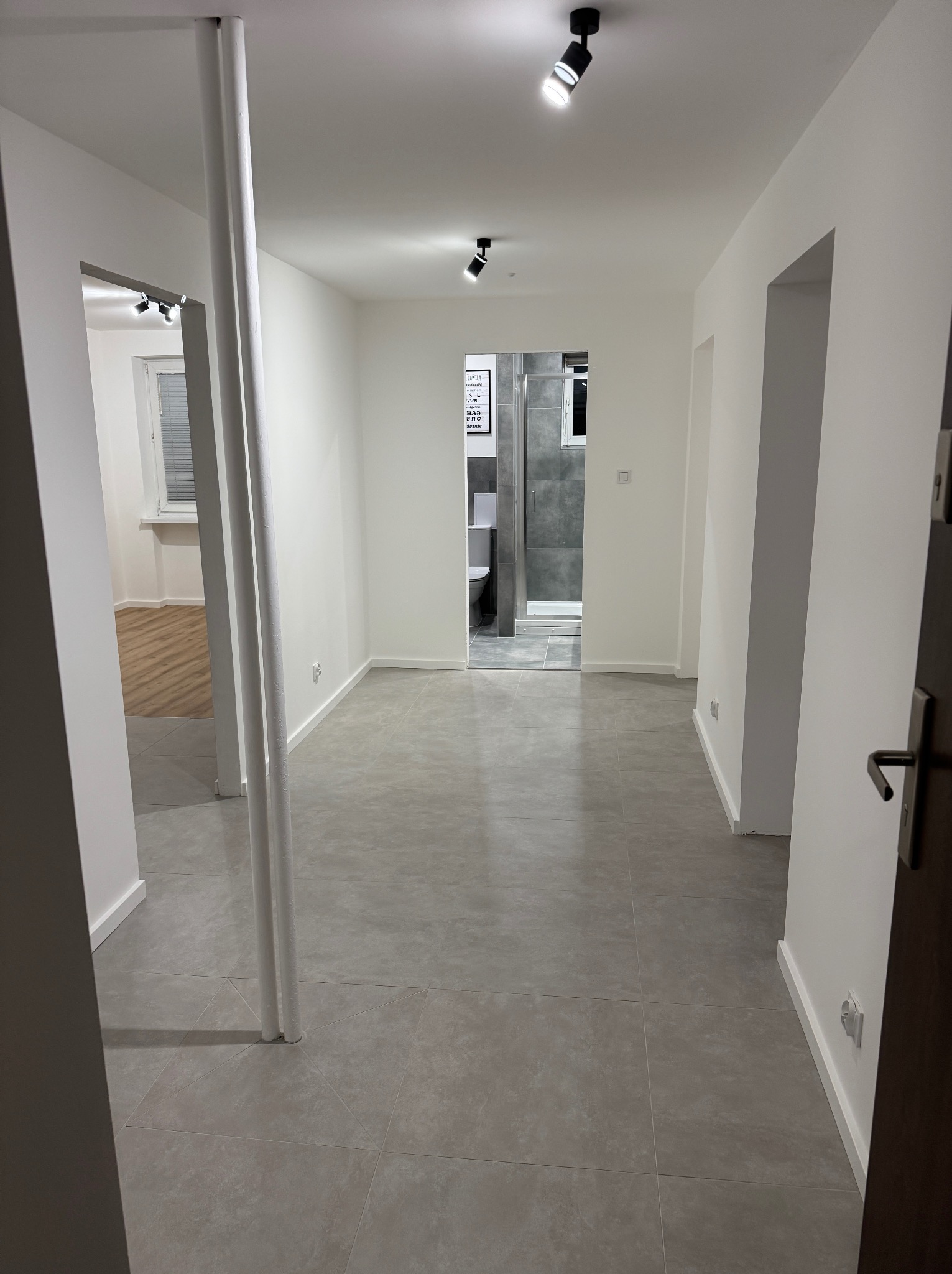 Wnętrze po remoncie: szare płytki podłogowe, białe ściany, widok na łazienkę z kabiną prysznicową. Minimalistyczny design, nowoczesne oświetlenie punktowe.